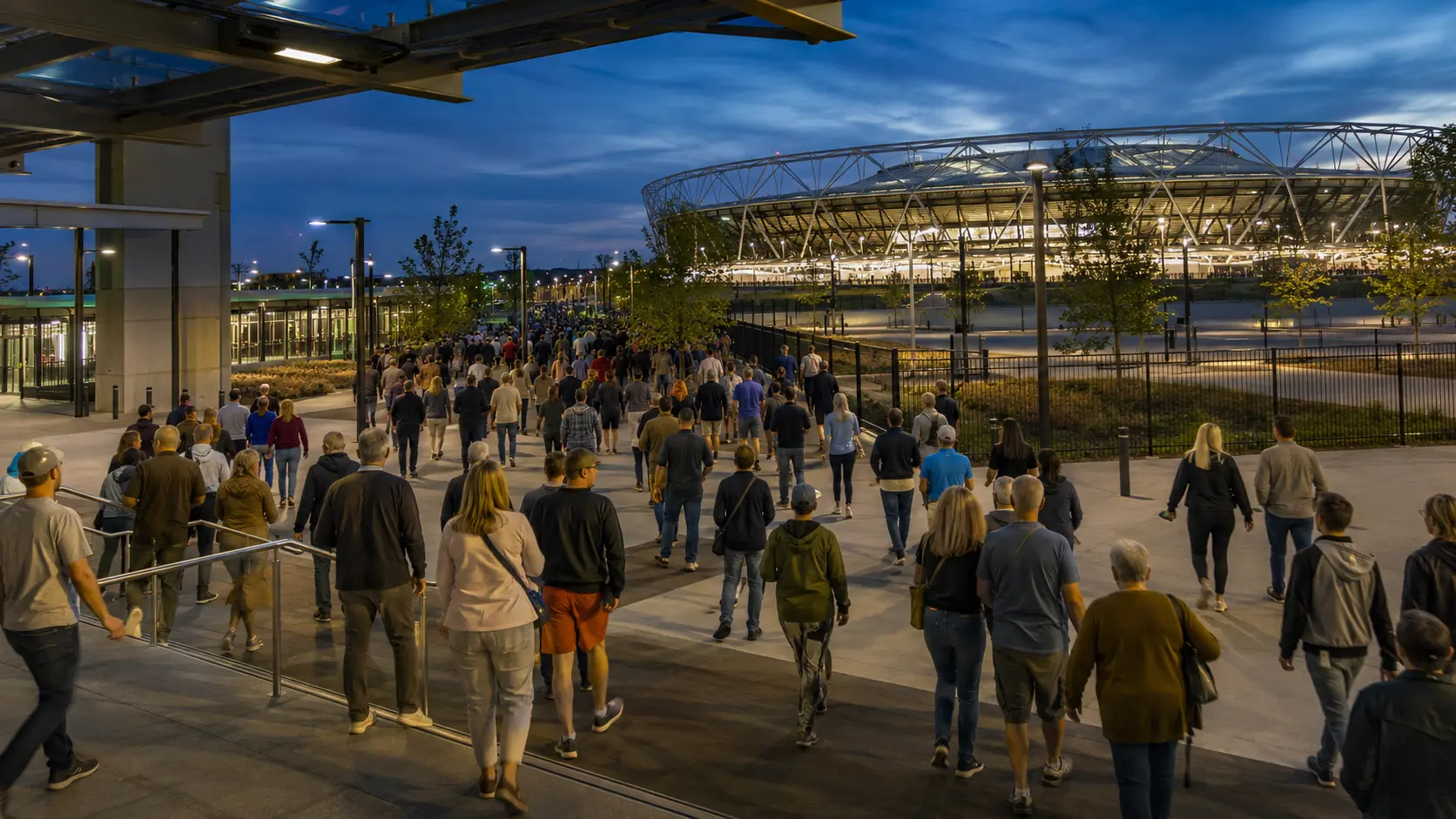 Eventgoers moving from a rail station toward a stadium at dusk